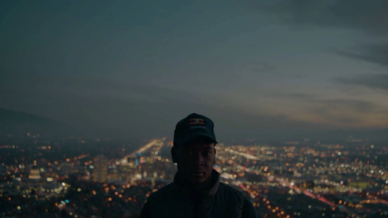 Cinematographer stands before city lights at dusk, wearing a hat with Uganda flag and text.