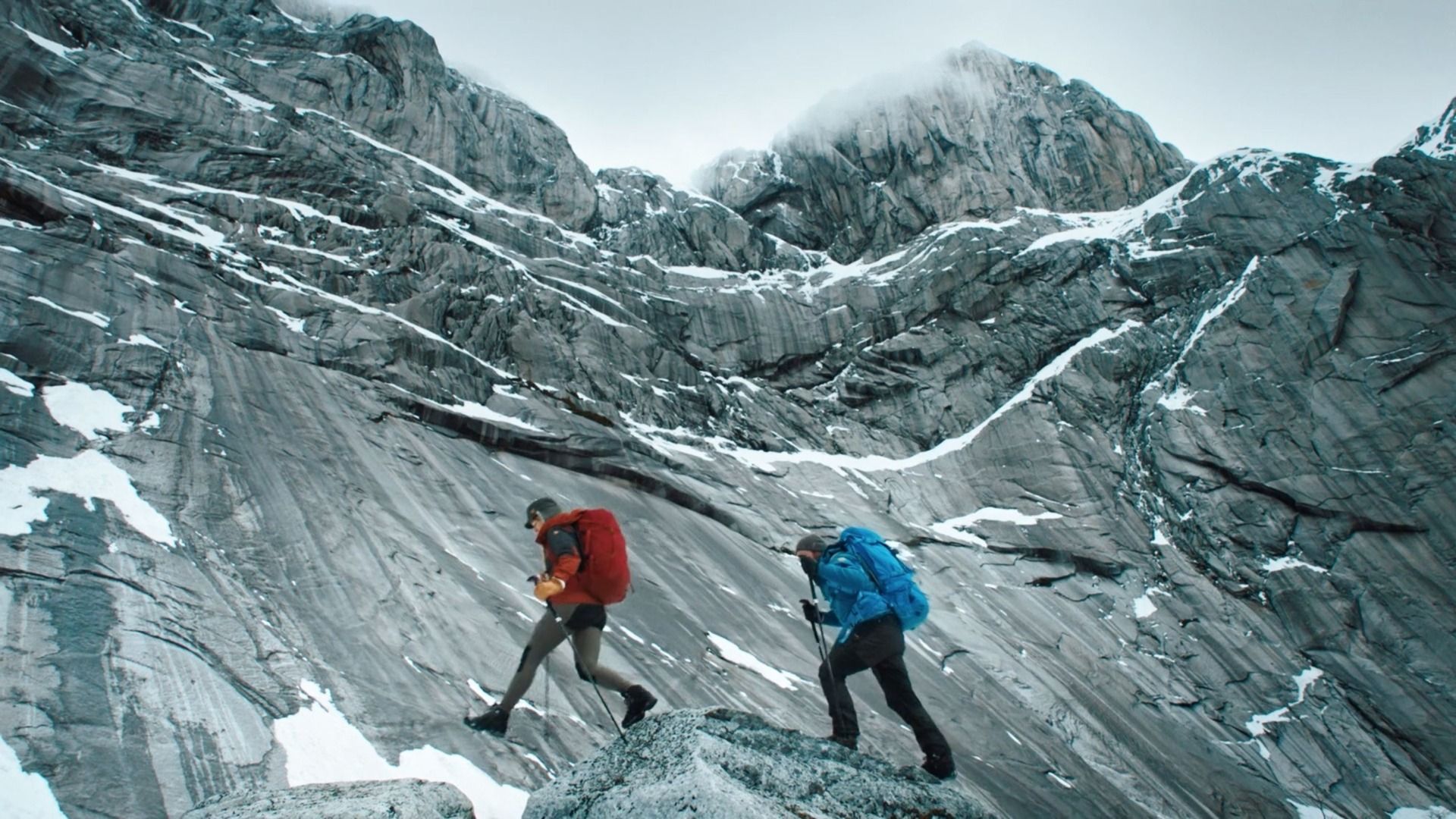 Two hikers with red and blue backpacks ascend a steep, rocky mountain slope with snow patches.