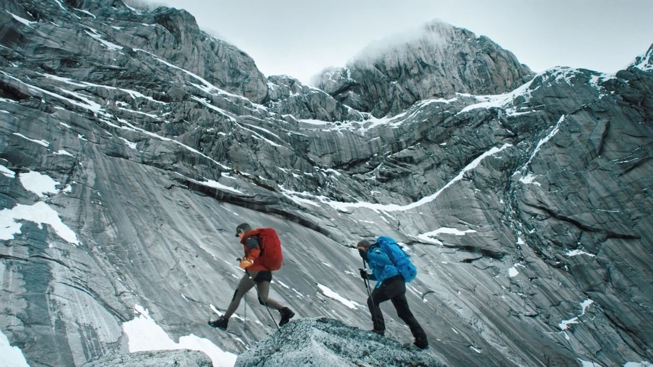 Two hikers with red and blue backpacks ascend a steep, rocky mountain slope with snow patches.