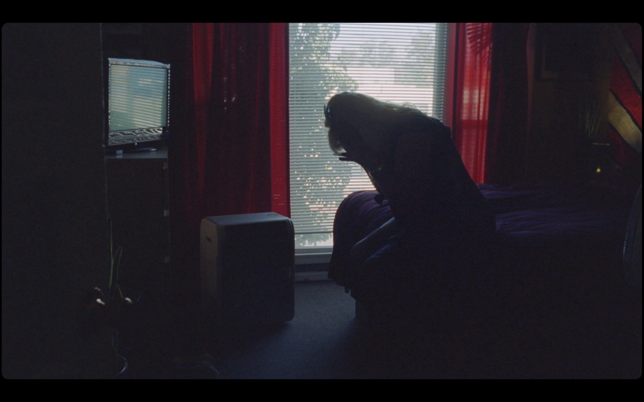 Silhouette of person sitting on bed in dimly lit room with window and red curtains