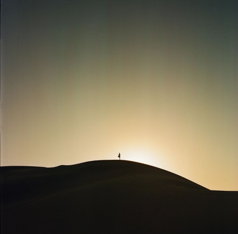 Silhouette of a lone hiker standing atop a desert dune against a glowing sunset sky.