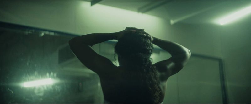 Back view of a female athlete with long curly hair, hands on head in a moody green-lit room.