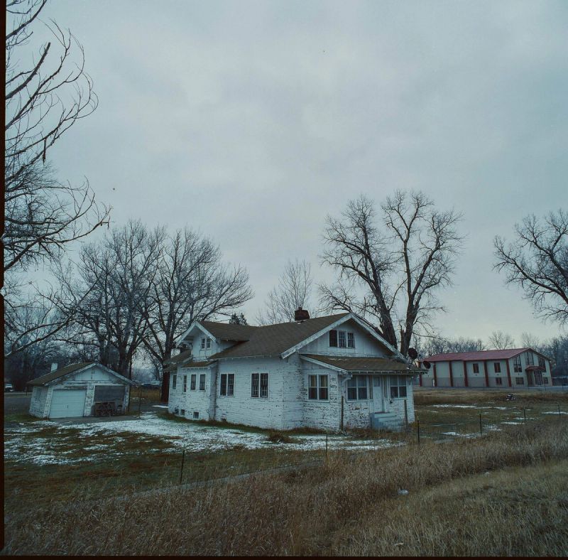 Weathered white farmhouse and garage in a snowy rural landscape under an overcast sky.