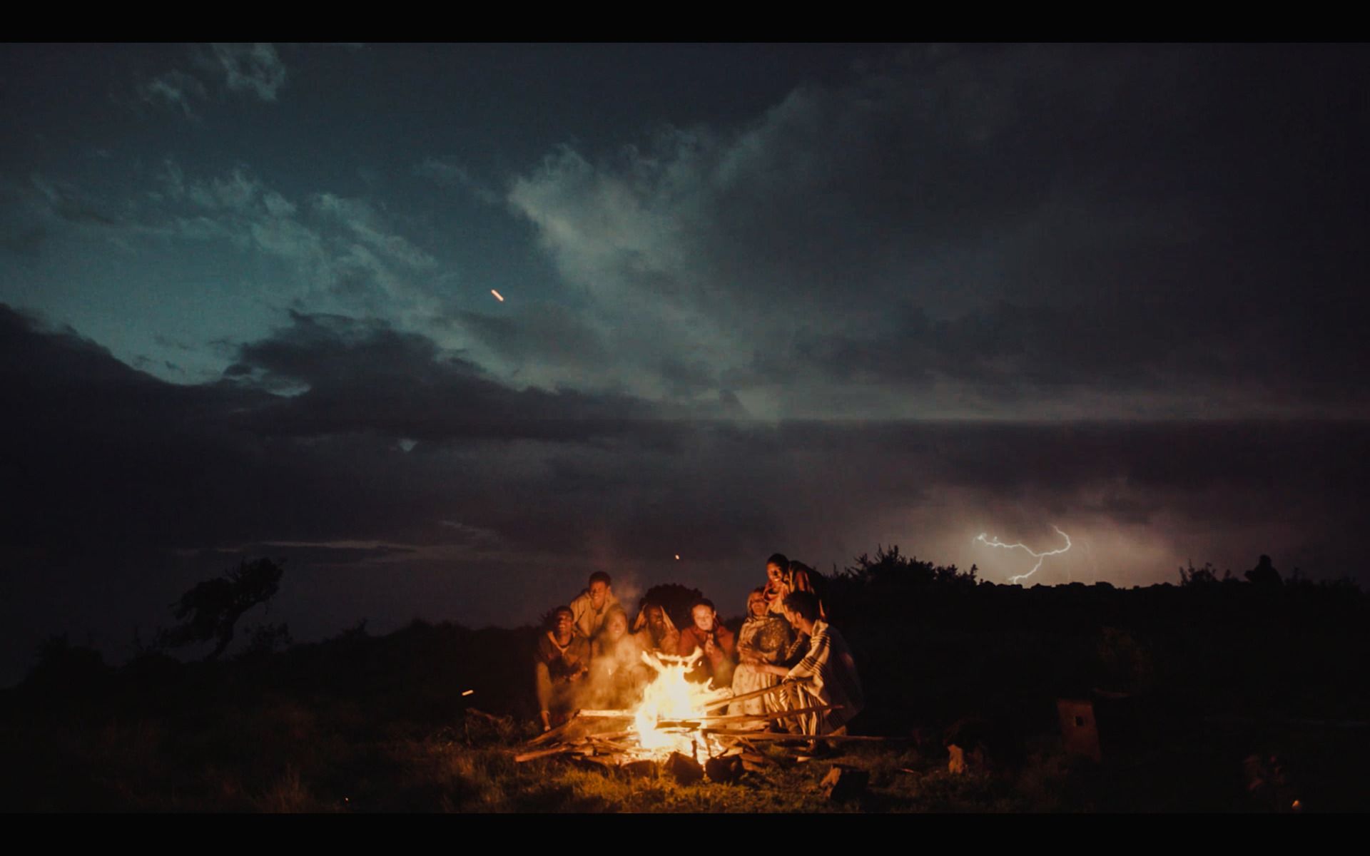 Group gathered around campfire under stormy night sky with lightning in distance