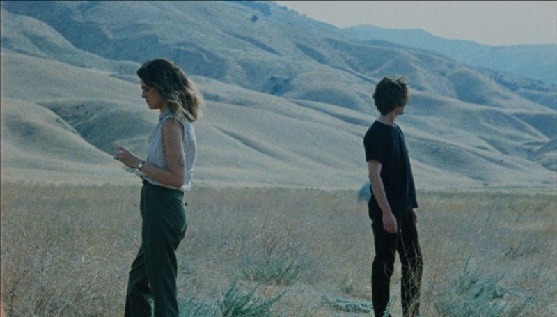 Two people stand apart in a dry, hilly landscape under soft light.