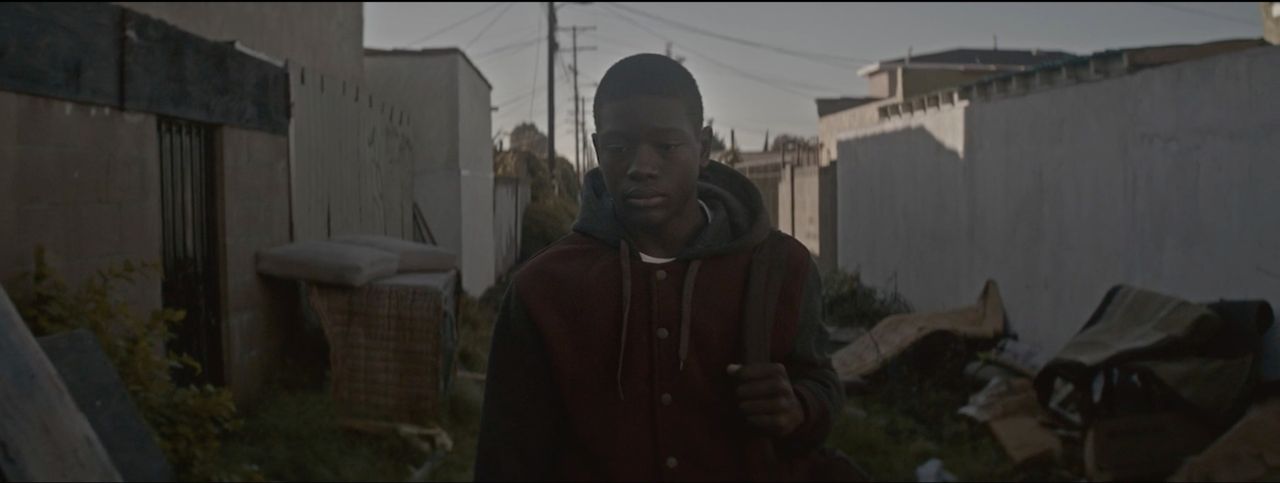 Young man in hoodie walking through narrow alleyway with buildings and debris