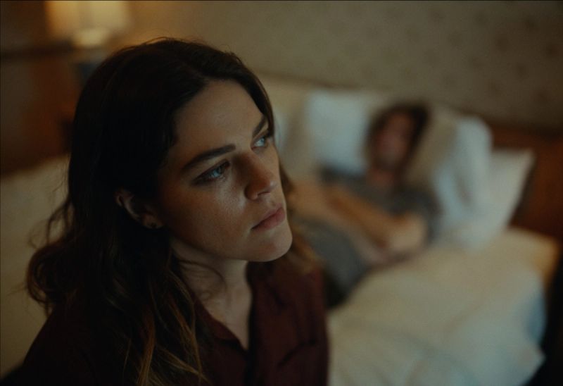 Woman in foreground looks thoughtfully while man sleeps in background on dimly lit bed