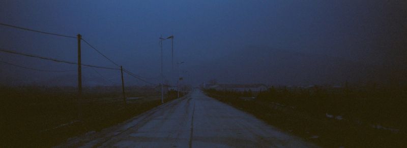 A desolate road stretching into the distance under a blue twilight sky, flanked by utility poles and streetlights.