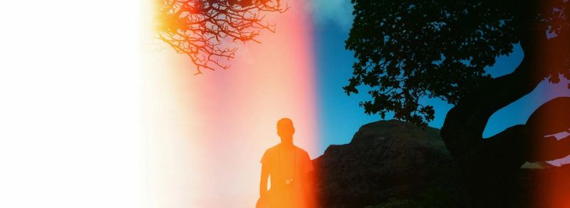 Silhouette of a man standing against a vibrant sunset sky with tree branches and rocky terrain.