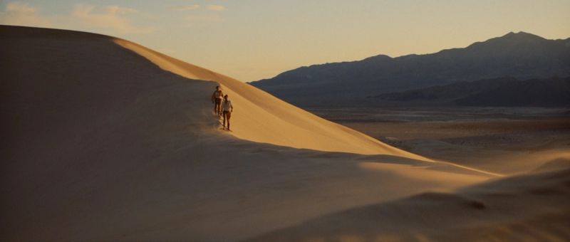 Two hikers ascend a sunlit sand dune in the desert at golden hour