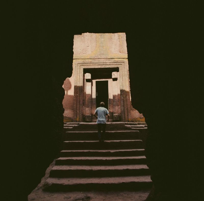A lone figure ascends stone steps toward a carved doorway in ancient ruins.