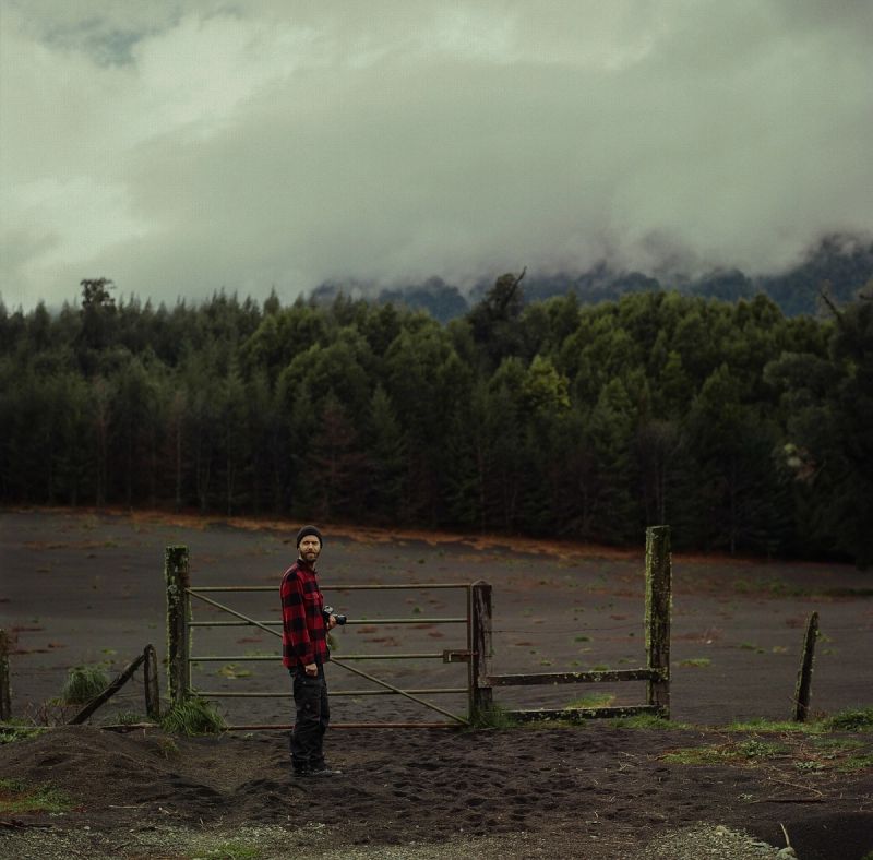 Cinematographer stands near a wooden gate in a dark field with misty mountains and forest background.