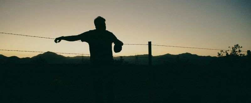 Silhouette of a figure holding a hat near a barbed wire fence with mountains in the distance at dusk.