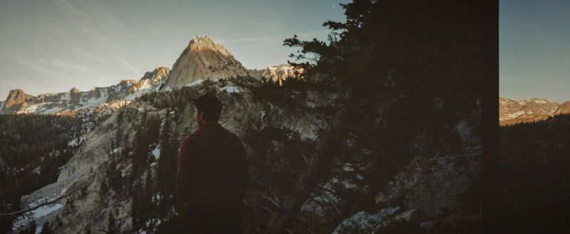 A lone figure overlooks rugged mountain peaks and pine trees in the Mammoth Mountains during golden hour.