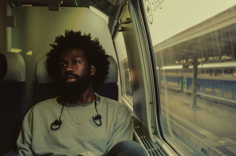 Man with afro sits on train seat looking out window at station platform, film photography.
