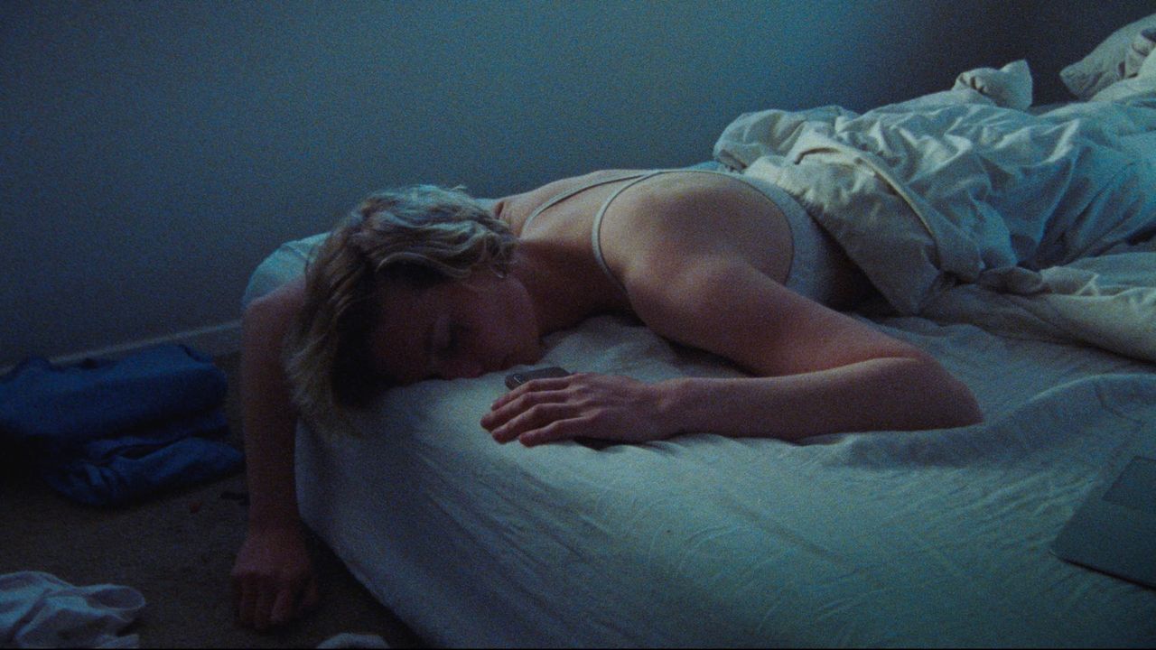 Woman sleeping on bed in dim blue-lit room, film photography still from Wet Nurse
