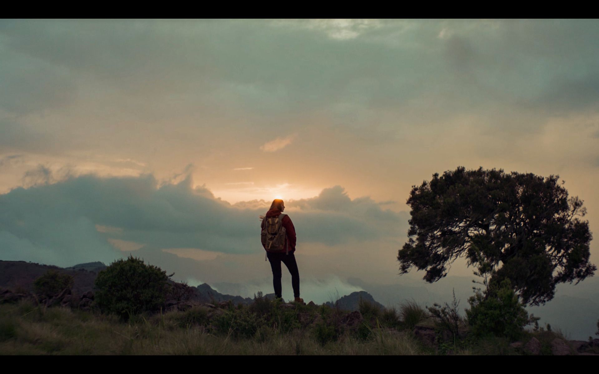 Hiker with backpack stands on mountain ridge at sunset, silhouetted against cloudy sky