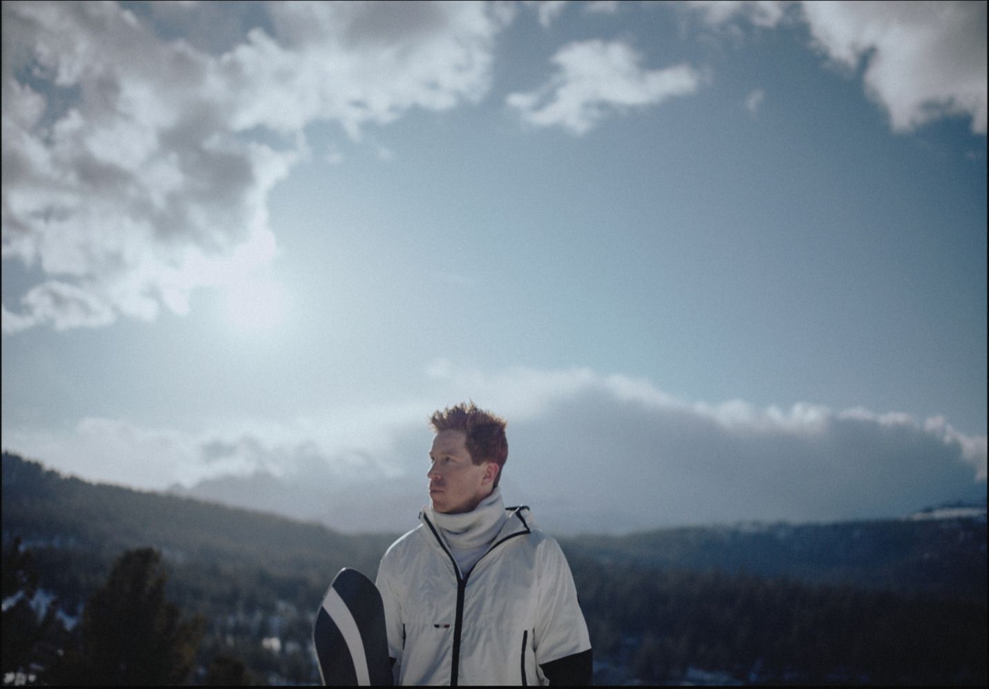 Snowboarder in white jacket stands against snowy mountain backdrop under cloudy sky
