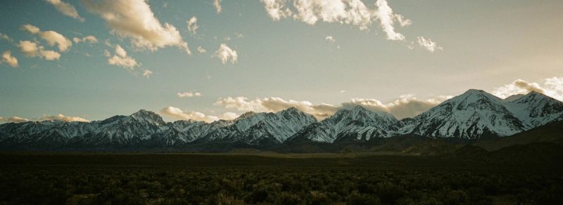 Snow-capped mountain peaks rise above a dark green valley under a cloudy sky.