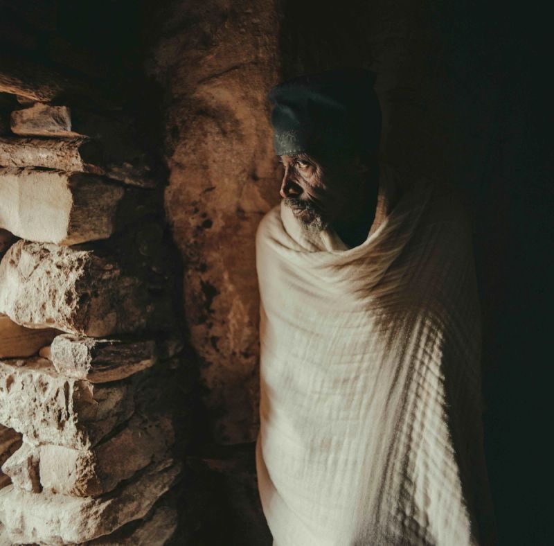 Cinematic portrait of an elder wrapped in white shawl inside a rustic stone structure.