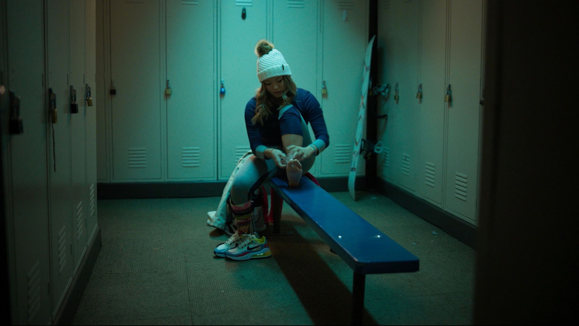 Chloe Kim tying her shoe in a locker room with a snowboard leaning against the wall.
