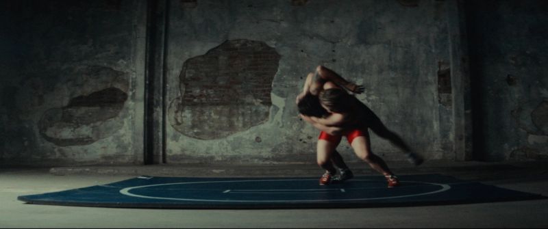 Two wrestlers grappling on a blue mat against a weathered, peeling concrete wall in an industrial space.