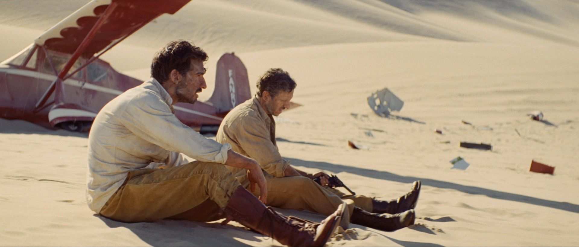 Two men sit in desert sand near a crashed red plane, cinematic film photography