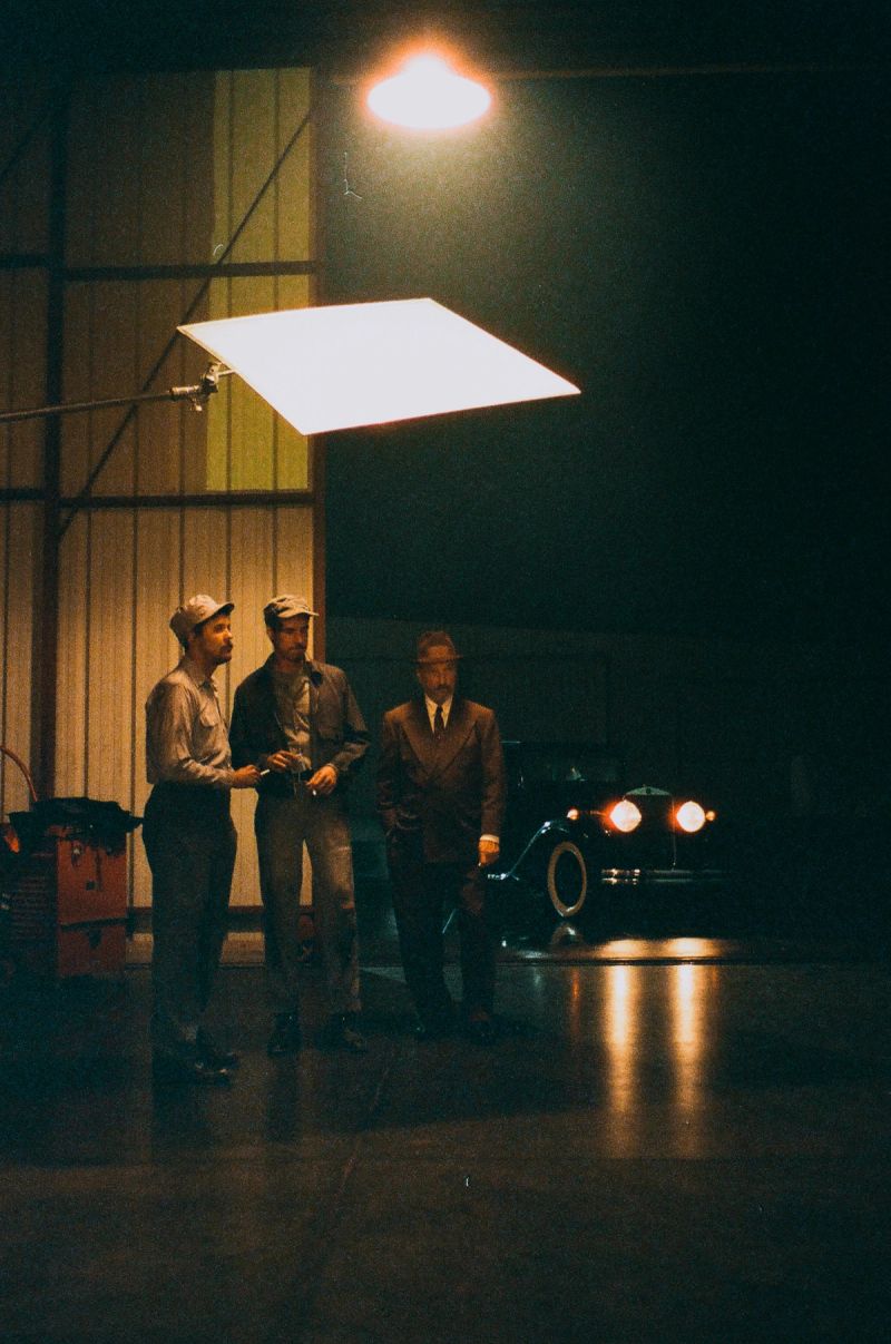 Three men on a dimly lit film set with vintage car and overhead lighting