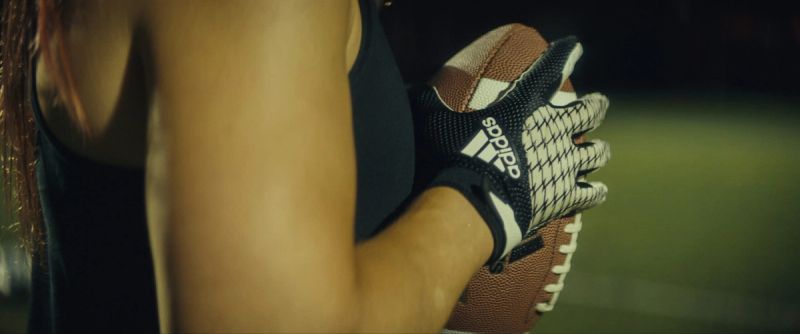 Close-up of a woman's arm holding an American football while wearing Adidas gloves.