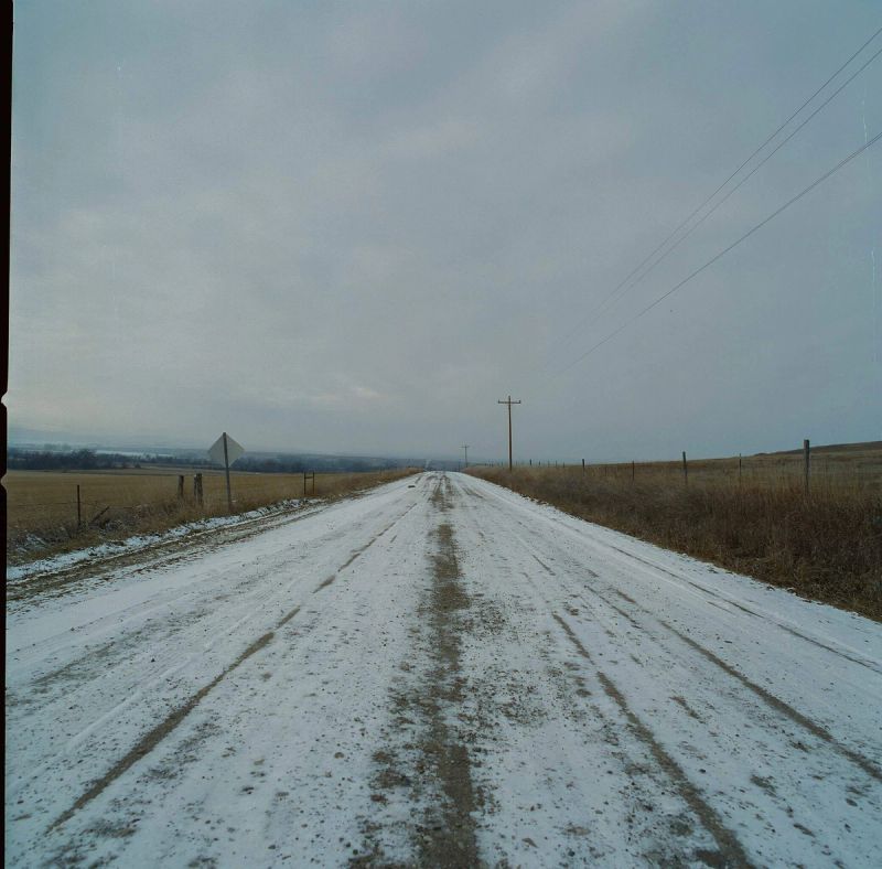 Snow-covered rural road stretching into overcast sky with power lines and fields