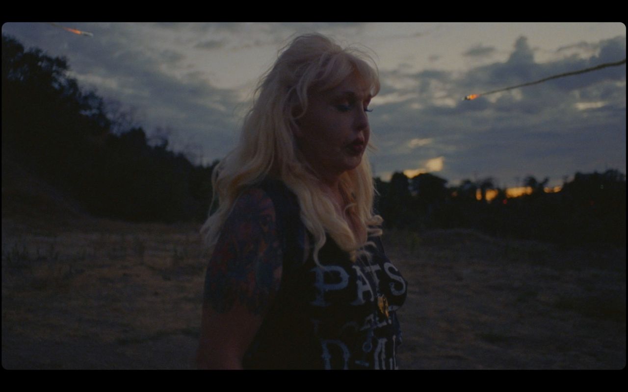 Blonde woman with tattoos and 'PATS' shirt in twilight desert landscape