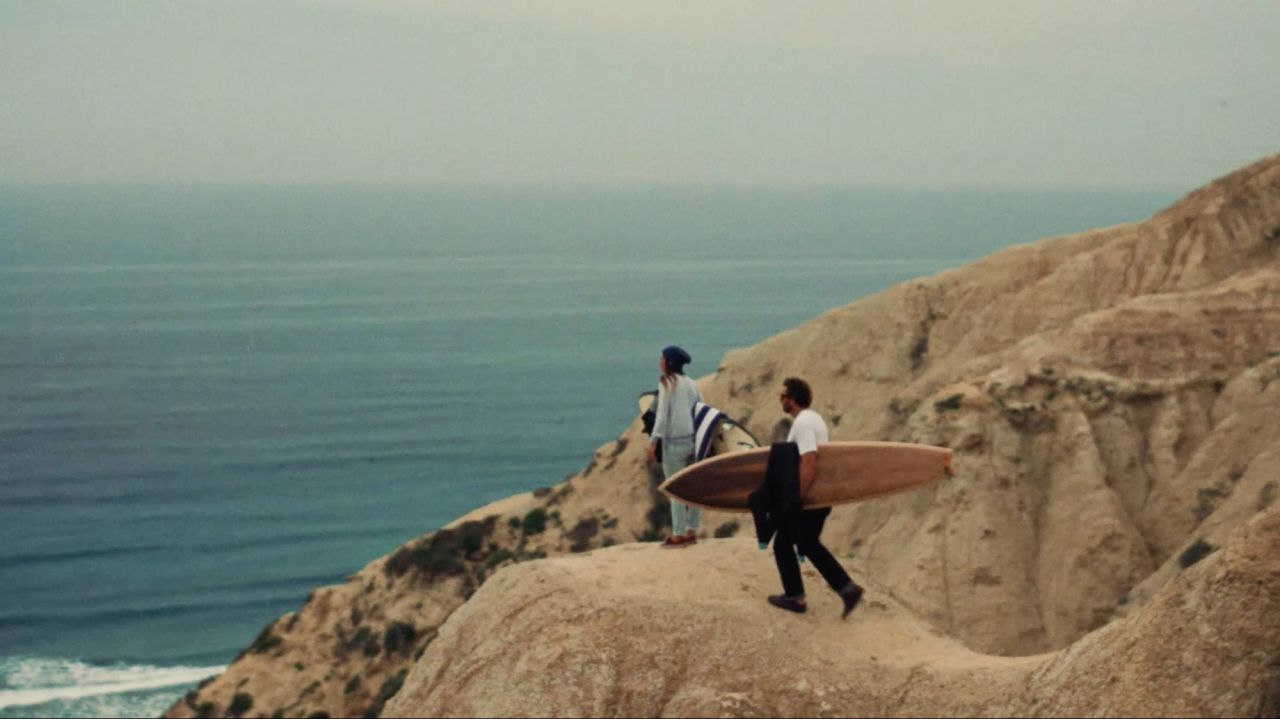 Two surfers carrying boards on a coastal cliff overlooking the ocean