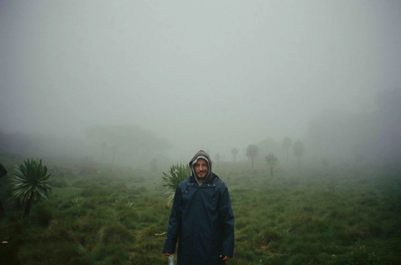 Man in hooded raincoat stands in misty green field with distant palm trees.