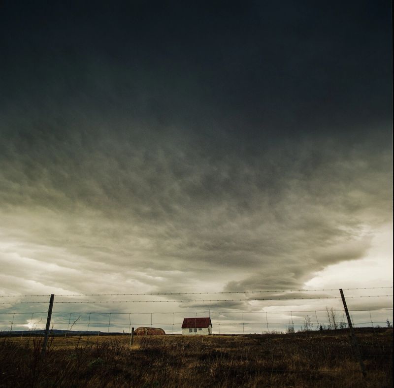 Solitary white house with red roof sits in a field under a dramatic, dark cloudy sky in Iceland.