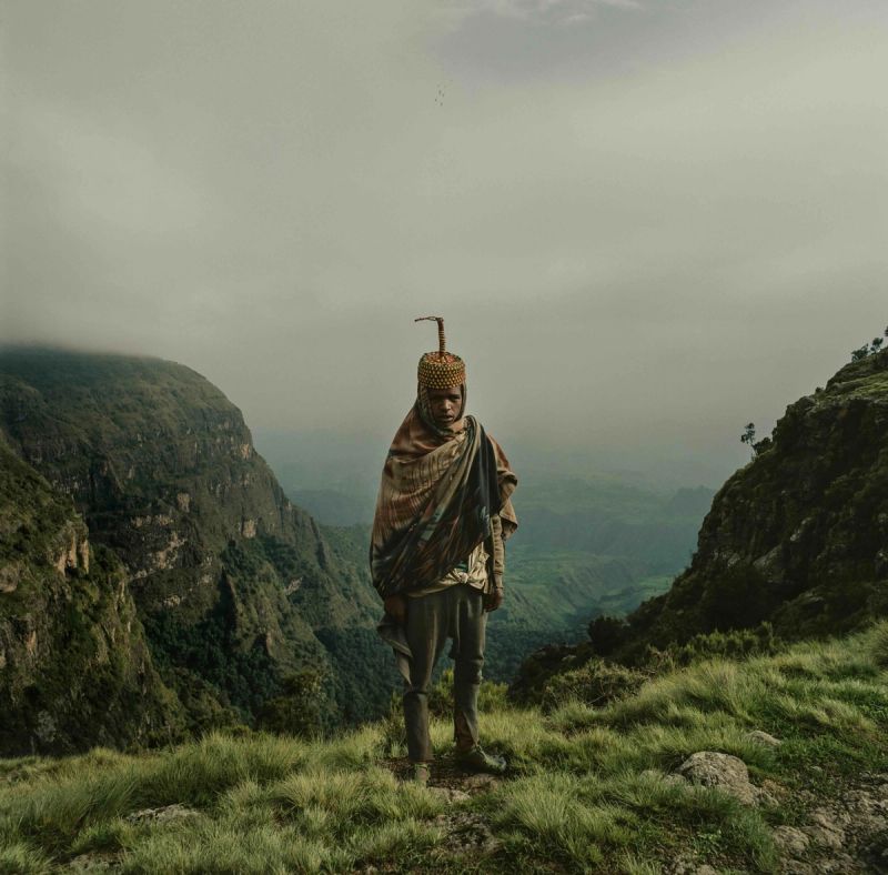 Ethiopian man in traditional headdress stands on mountain ridge with misty valley below