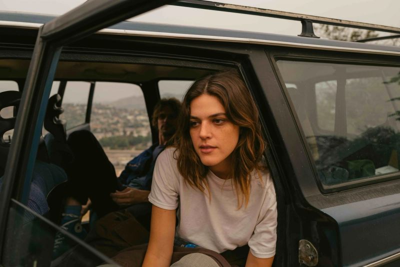 Woman in white t-shirt looking out from open car door with hazy mountain landscape behind.