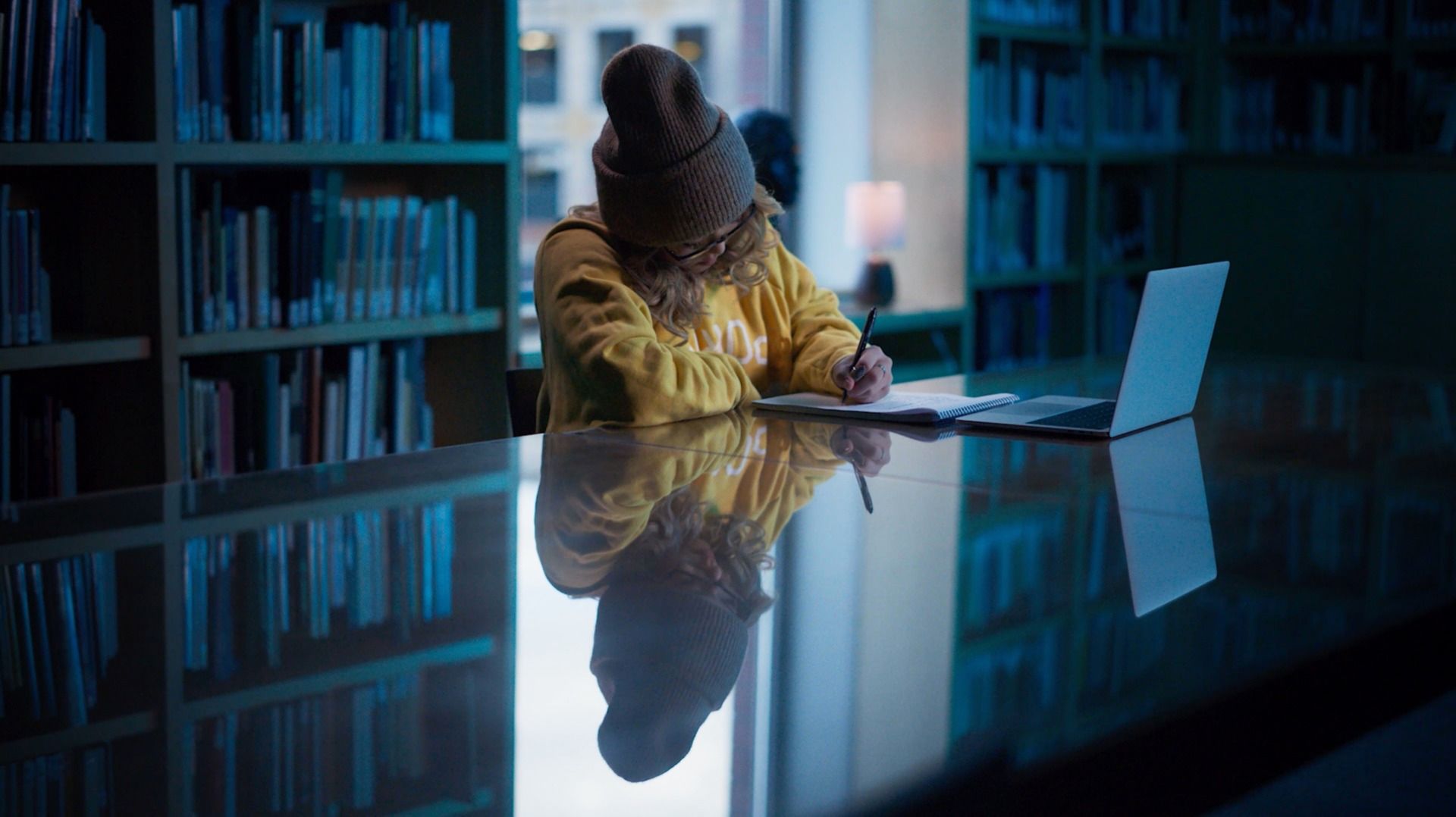 Chloe Kim studies at library desk with laptop and notebook, wearing yellow hoodie.