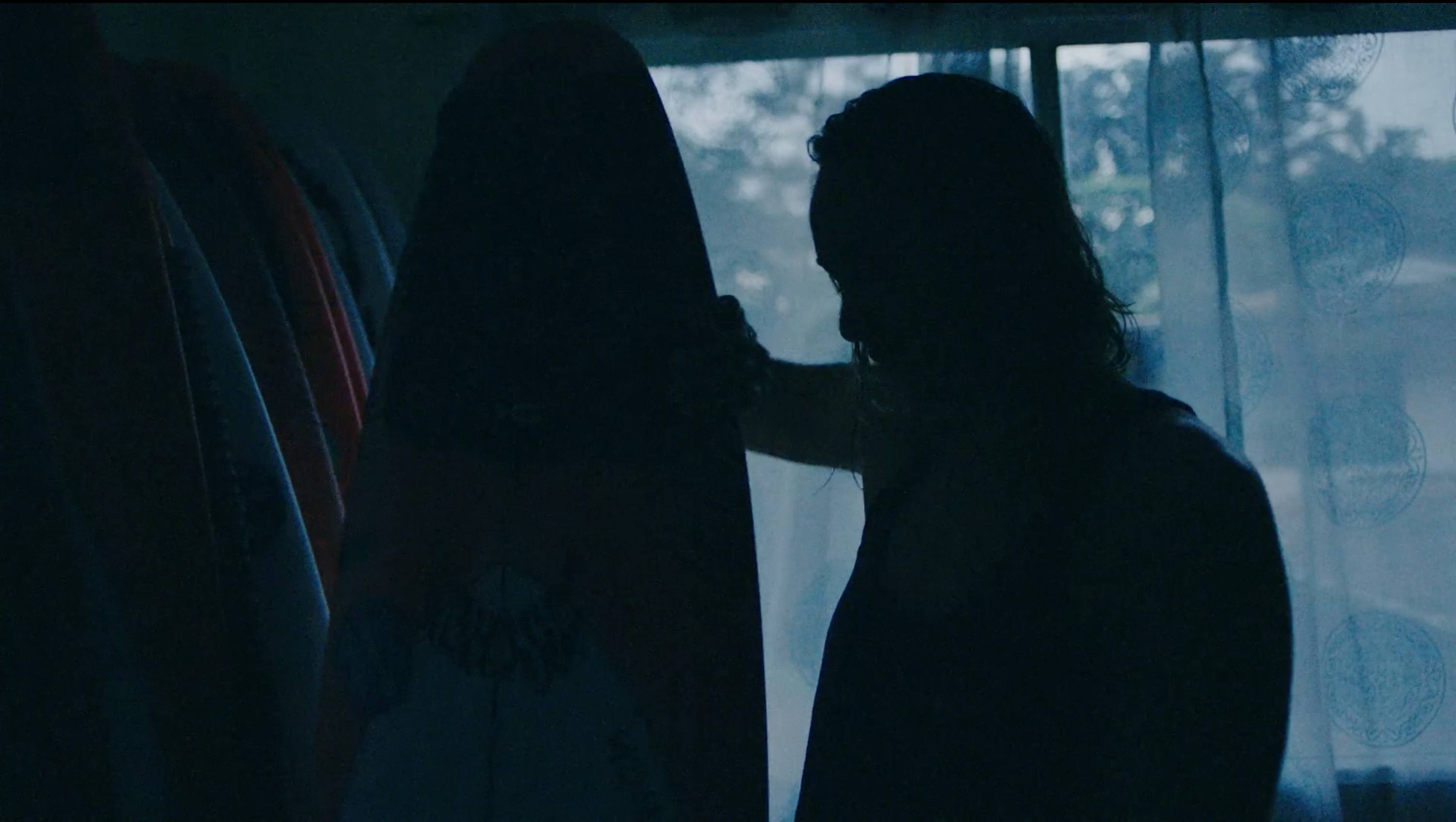 Silhouette of person holding surfboard in dimly lit room with window and patterned curtains