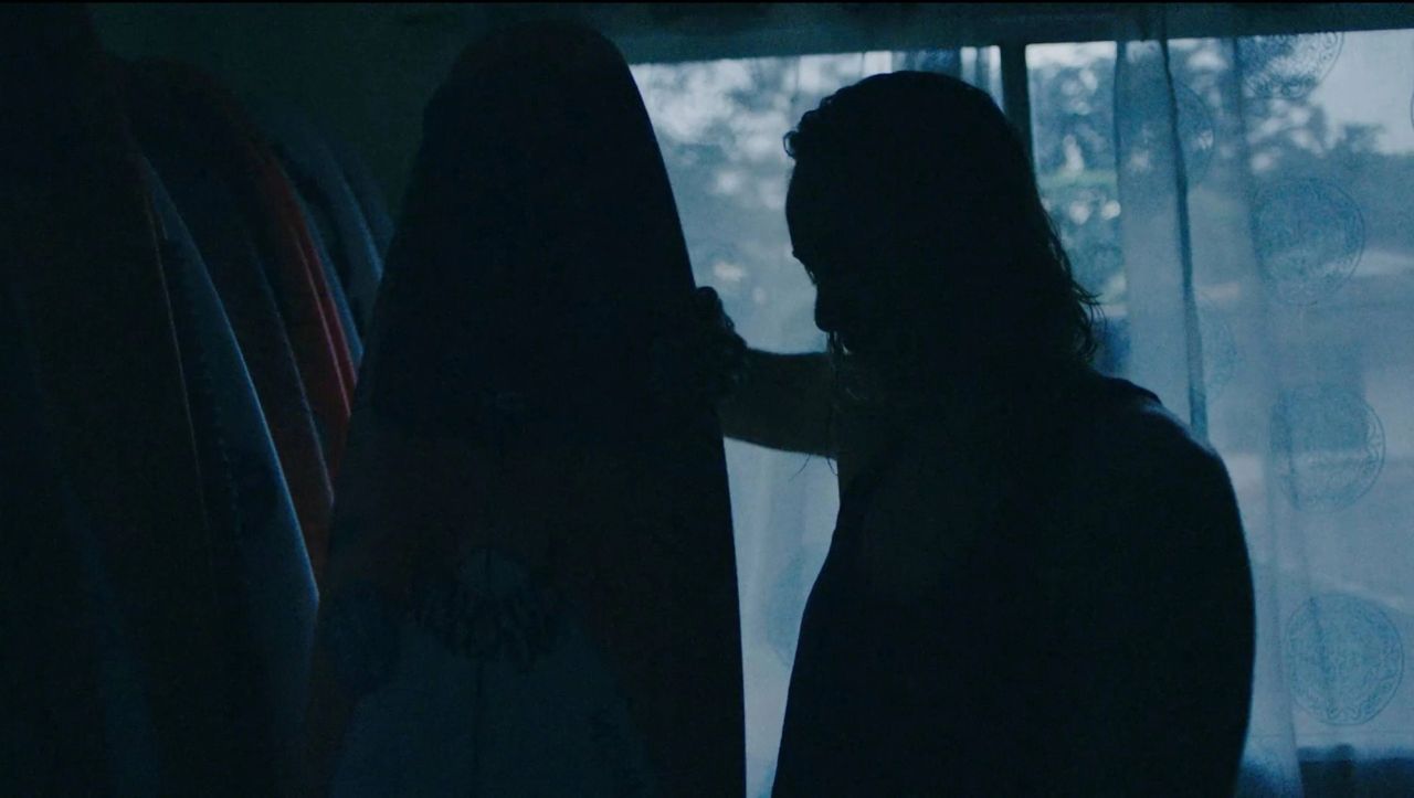 Silhouette of person holding surfboard in dimly lit room with window and patterned curtains
