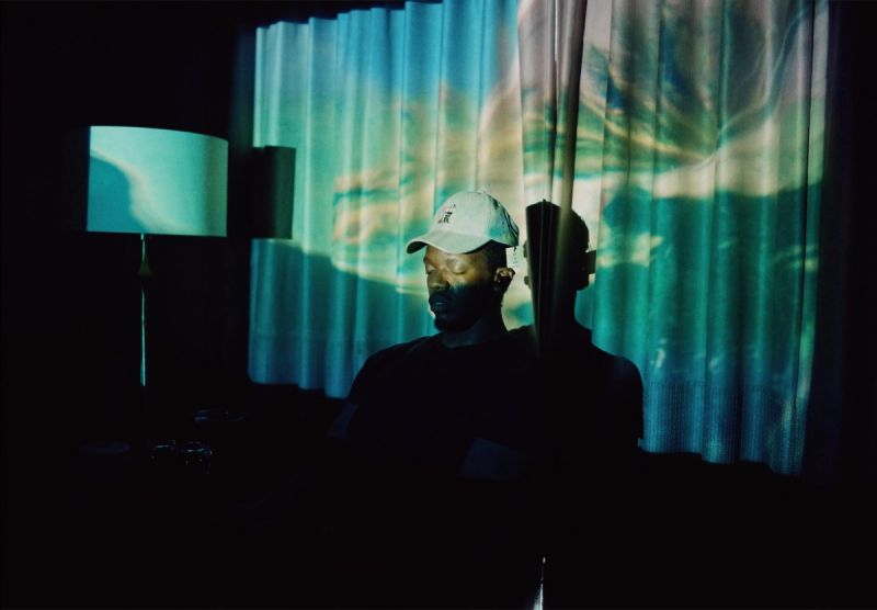 Reggie Jackson sits contemplatively in a dark room with teal light projected on curtains, captured on Kodak 16mm film.