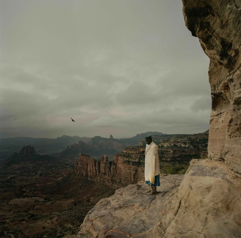 A figure in white robes stands on a rocky cliff edge overlooking vast canyon landscapes under an overcast sky.