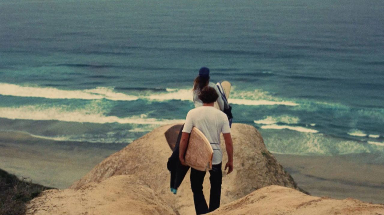 Two surfers with boards walk down a sandy cliff toward the ocean waves.