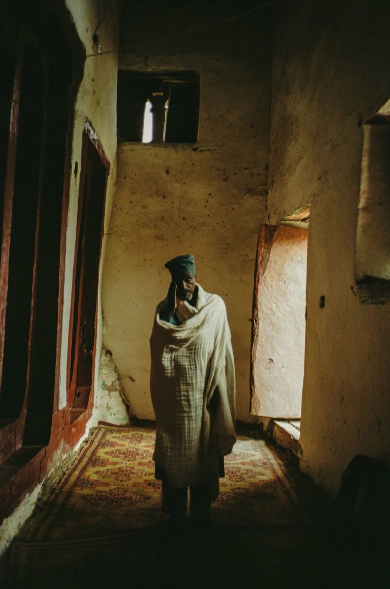 Man wrapped in shawl stands on patterned rug in rustic interior hallway with window light.