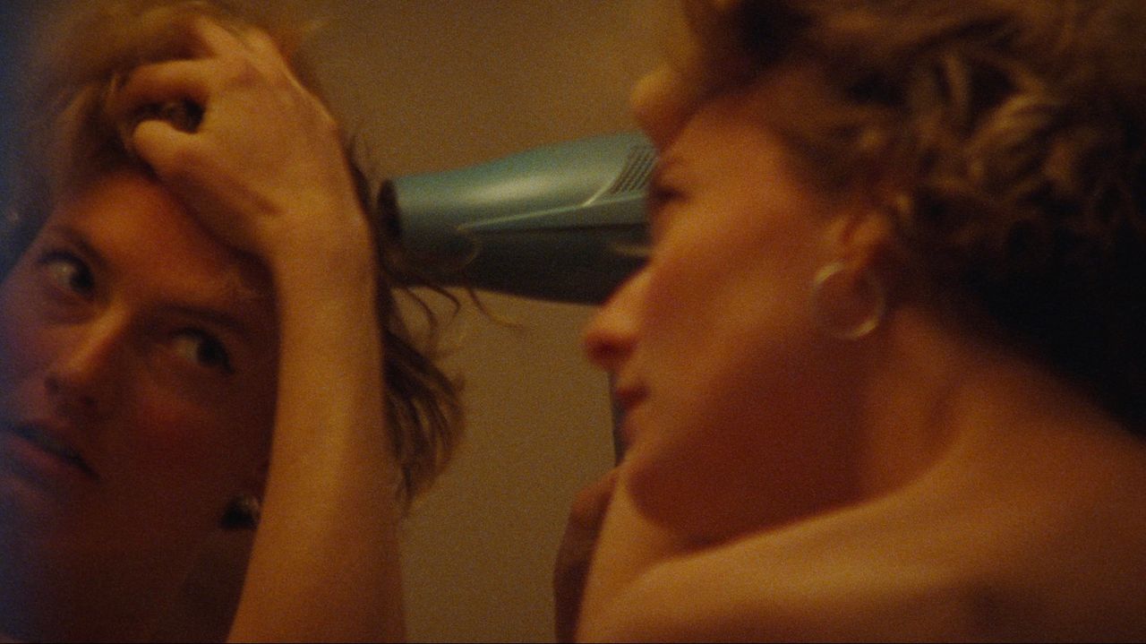 Woman blow-drying curly hair in warm-toned bathroom mirror reflection