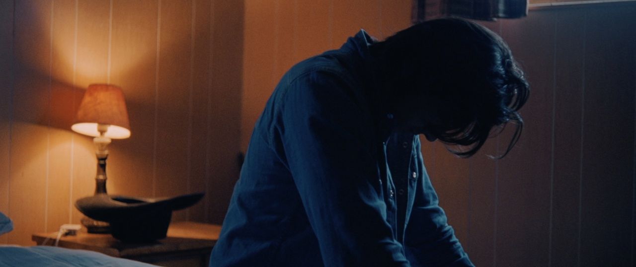 Person in dimly lit room with lamp and wooden wall, head bowed