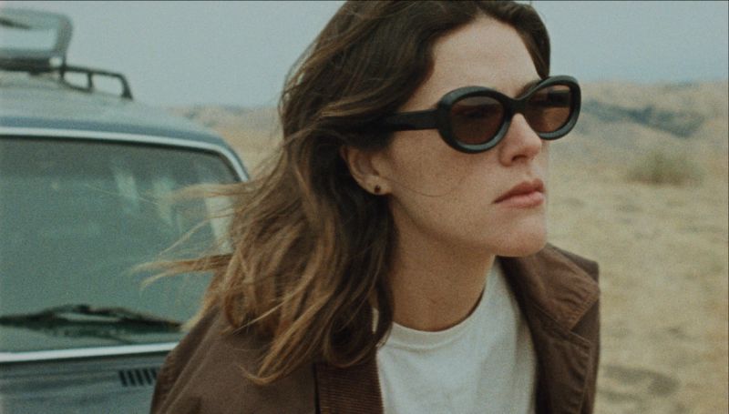 Woman in sunglasses and brown jacket stands near vintage car in desert landscape