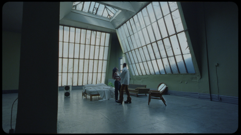 Two people stand in a sunlit, dilapidated room with large broken windows and vintage furniture.