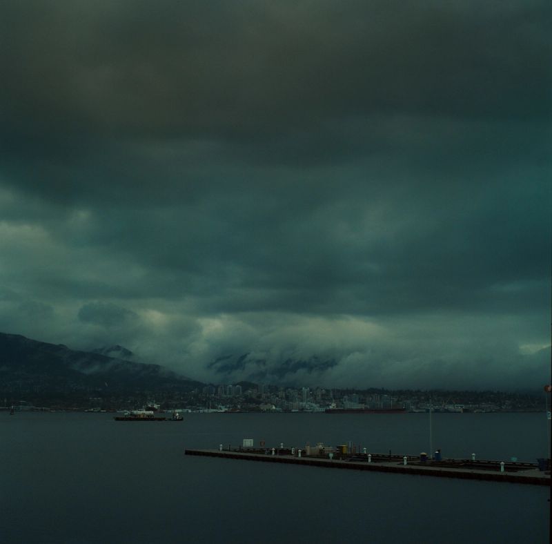 Dark storm clouds over a coastal city skyline with mountains and harbor in the background