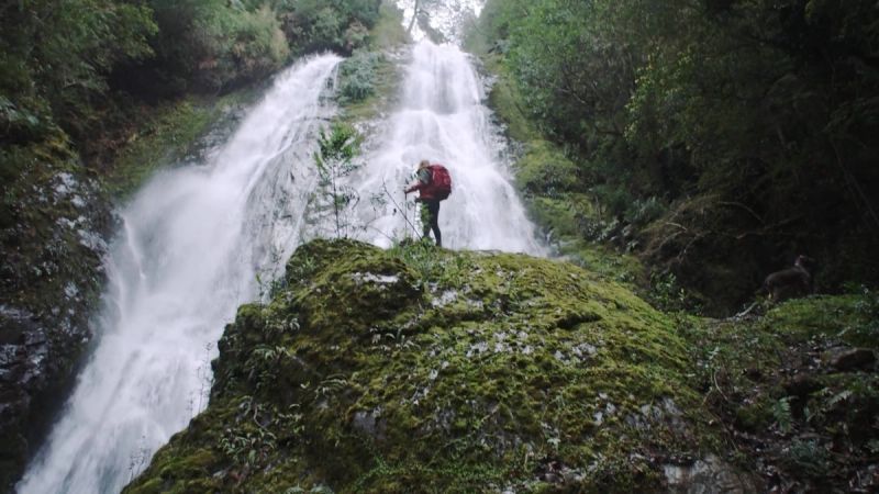 Cinematographer captures hiker with red backpack standing on mossy rock before cascading waterfall.