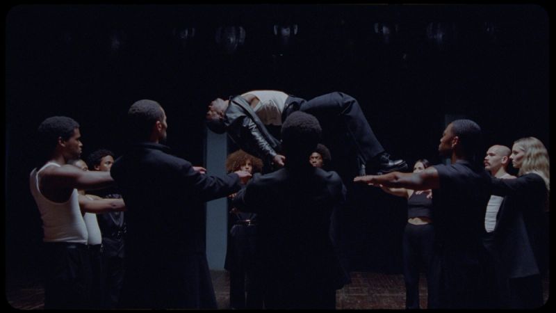 Dancers in dark suits lift a performer mid-air in dramatic choreography